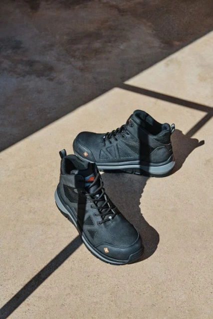 Pair of black leather work boots are placed in direct sunlight. The composition is a product shot on concrete flooring.