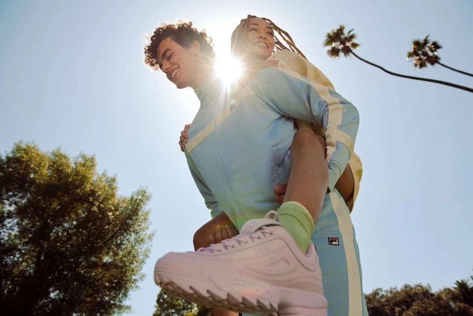 Smiling couple in Fila apparel: man giving woman a piggyback ride outdoors under sunny sky with palm trees and pink sneakers.