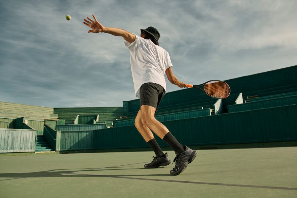 Man in a bucket hat and black tennis attire throws a tennis ball on a green tennis court, preparing to serve, with stadium seating behind him.