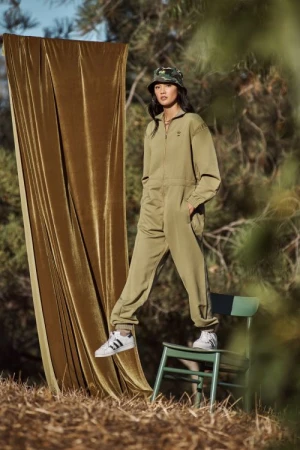 A woman in an olive green jumpsuit and camouflage bucket hat poses on a chair in an outdoor setting with a draped curtain.