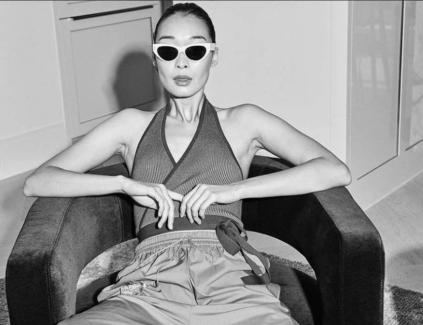Black and white portrait of a stylish adult woman sitting in a chair wearing sunglasses, a halter top, and pants in an indoor setting.