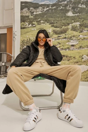 Woman in sunglasses and puffer jacket poses in a chair in a photo studio with a mountain scene backdrop, wearing tan sweat suit and sneakers.