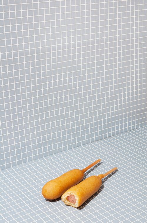 Two corn dogs are arranged on a blue-grey checkered surface with matching wall in a brightly lit studio setting, food photography.