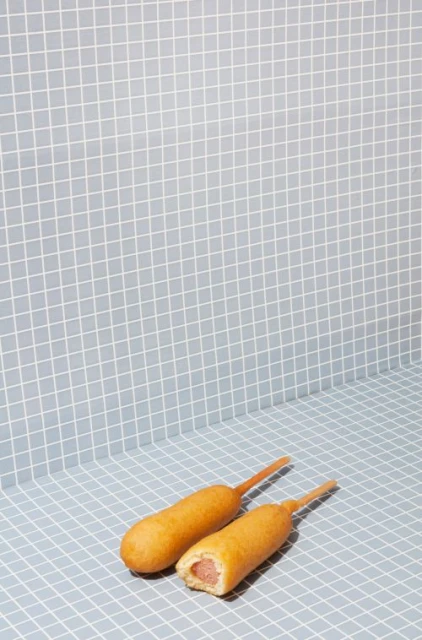 Two corn dogs are arranged on a blue-grey checkered surface with matching wall in a brightly lit studio setting, food photography.