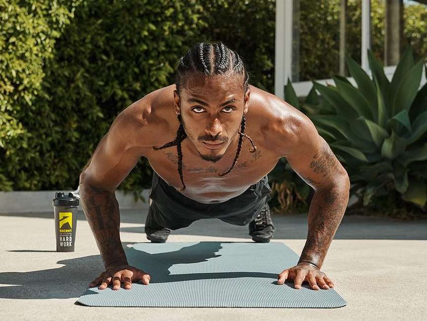 A man with braids is outdoors doing push-ups on a gray mat, with a shaker bottle nearby and green foliage visible in the background.