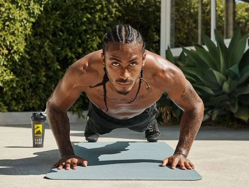 A man with braids is outdoors doing push-ups on a gray mat, with a shaker bottle nearby and green foliage visible in the background.