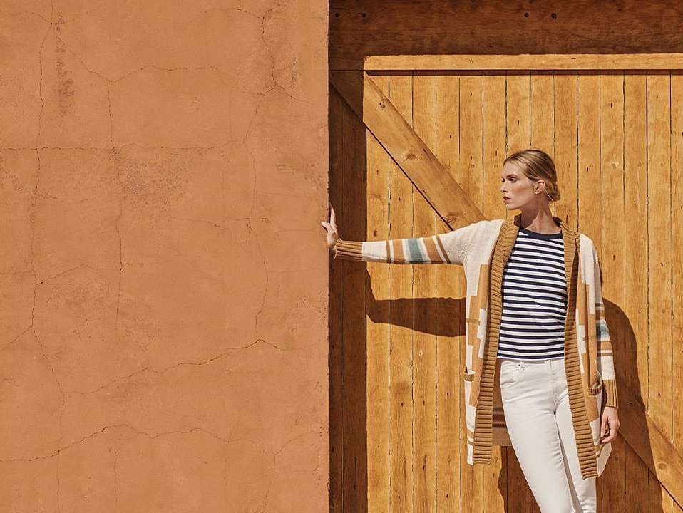 Adult woman in striped shirt and cardigan poses near orange wall and wooden door in natural light, fashion photography.