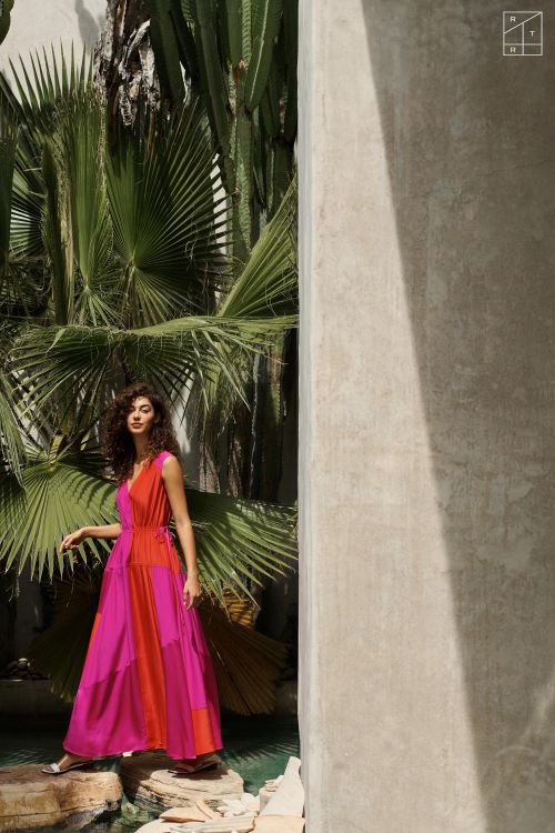 Woman in a bright pink and orange dress poses on rocks near water with tropical plants and white concrete wall in the background.
