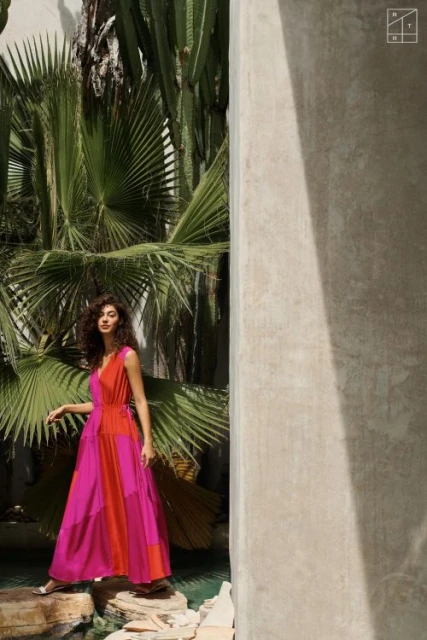 Woman in a bright pink and orange dress poses on rocks near water with tropical plants and white concrete wall in the background.