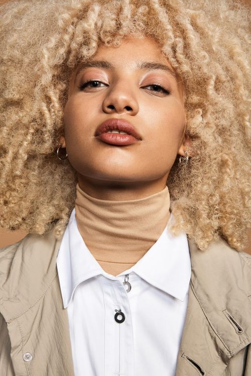 Close-up portrait of a stylish adult female with blonde curly hair, fair skin, neutral facial expression, and fashionable jacket posing indoors.