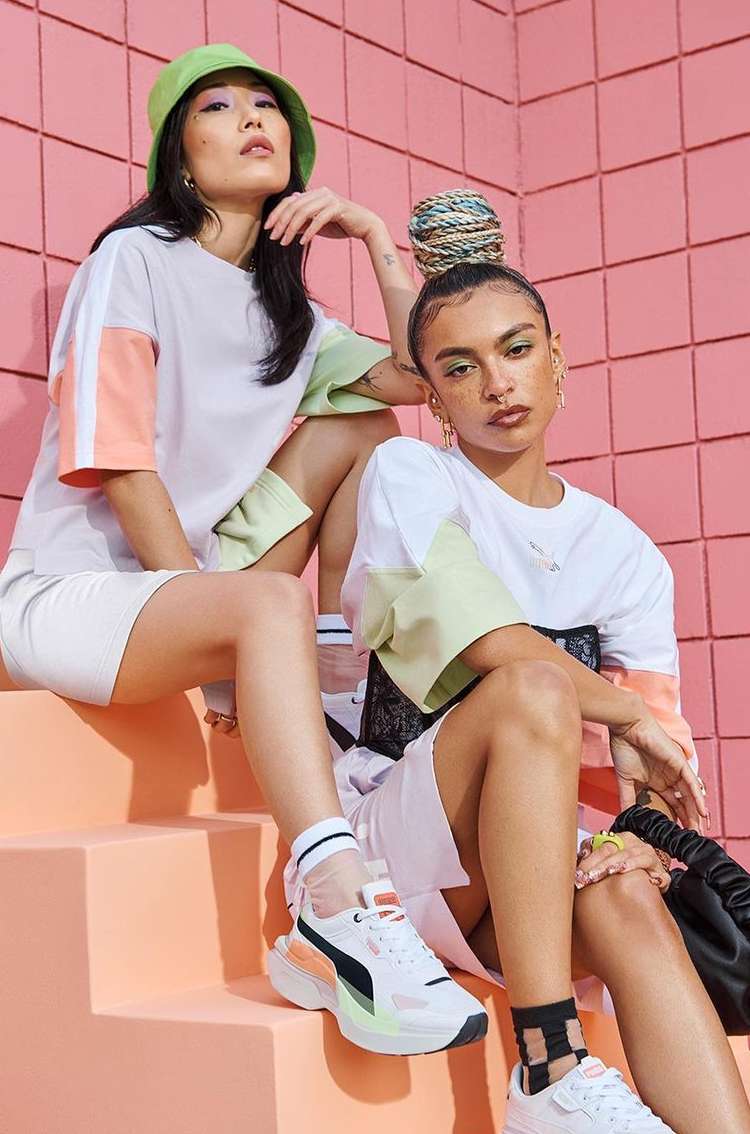Two young adult fashion models wear coordinating white t-shirts and athletic shorts while posing against a pink tiled wall.