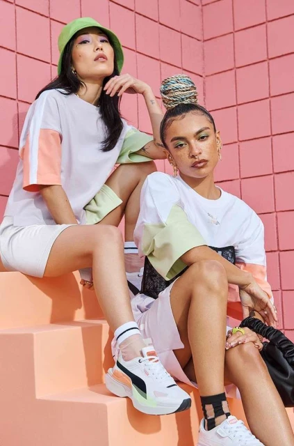 Two young adult fashion models wear coordinating white t-shirts and athletic shorts while posing against a pink tiled wall.