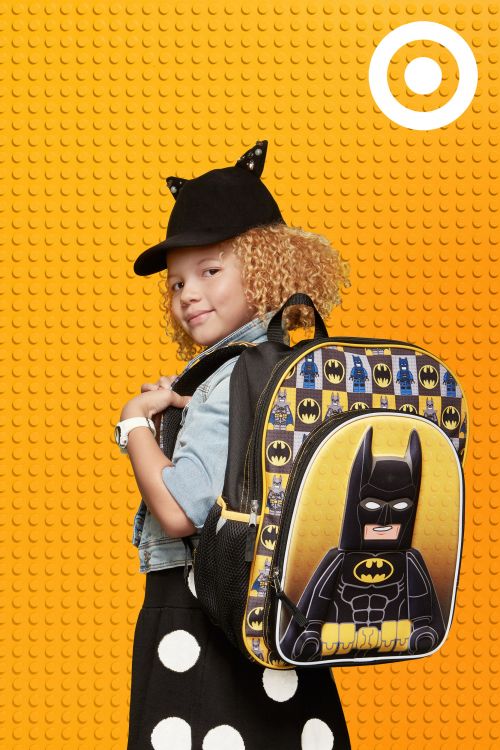 Young child models a Lego Batman backpack in front of a brick-patterned yellow background, accessorized with a cat-eared hat.