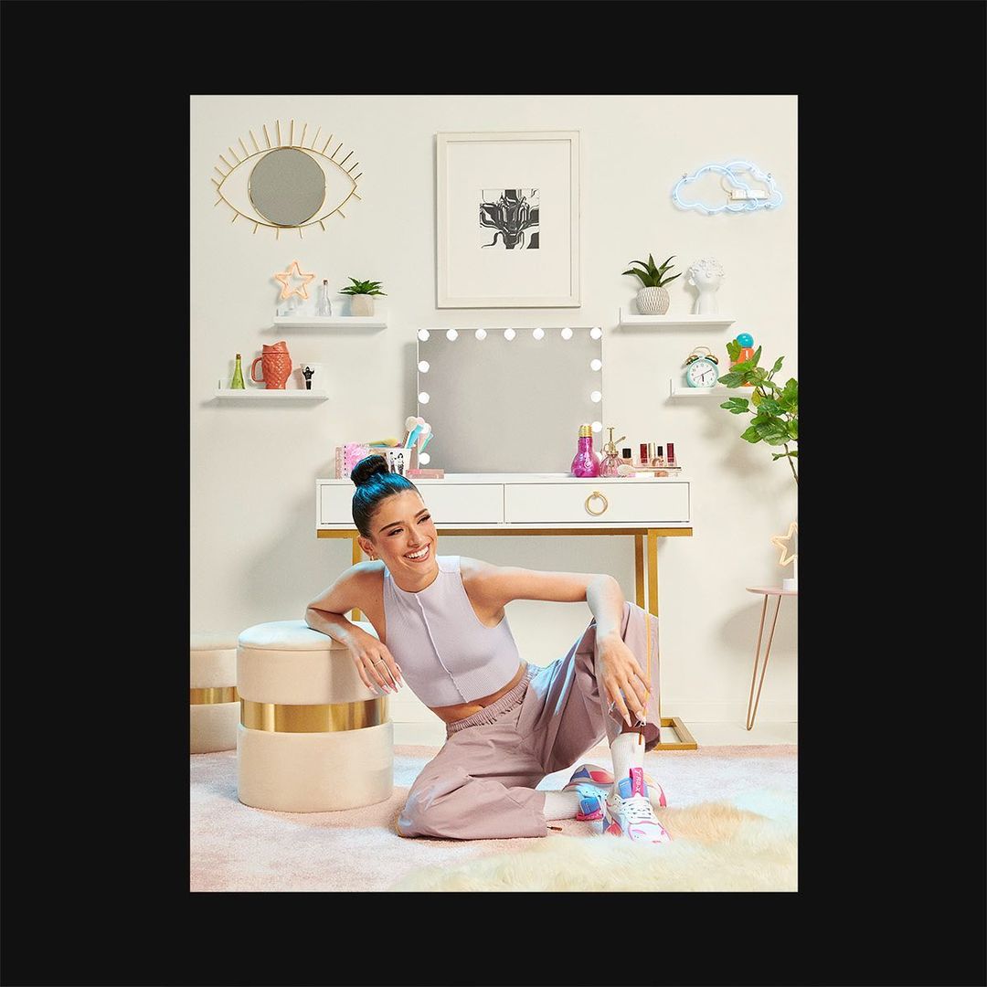 Smiling woman in a lavender top poses with her leg up in front of a white vanity in an indoor setting with neutral decor.