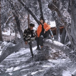 Two models sitting on a large snowy tree branch in a forest, wearing an orange puffer jacket and a camo jacket by Molly Cranna