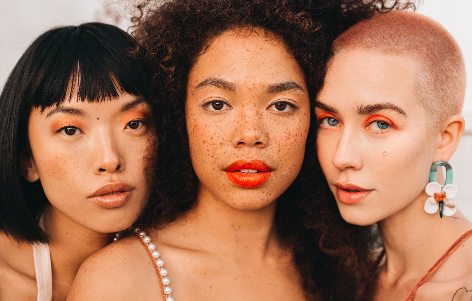 Close-up of three diverse women with colorful eye makeup and unique hairstyles posing in a brightly lit, minimalist indoor setting.