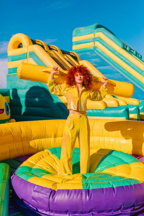 A woman with red curly hair poses on a large colorful inflatable, wearing a matching yellow jacket and pants under a bright blue sky.