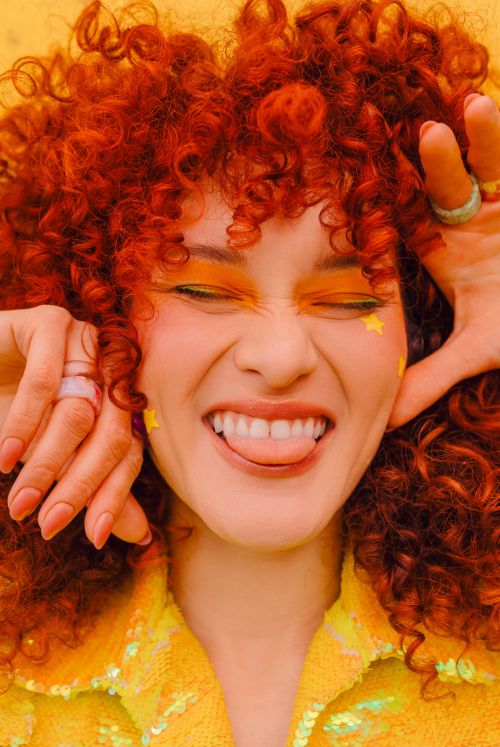 Smiling woman with curly red hair, yellow eyeshadow, star stickers, and a sparkly yellow jacket poses against a bright yellow backdrop.