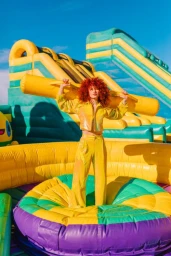 Woman with voluminous red hair wearing a yellow iridescent jacket and matching trousers posing on an inflatable playground structure