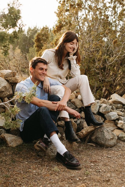 A couple poses outdoors near greenery; the man is seated while the woman sits behind him with her hand on his shoulder.