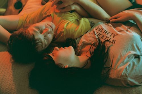 Two people lying on a bed, viewed from an overhead angle, captured with soft, warm lighting that highlights skin tones and textures.