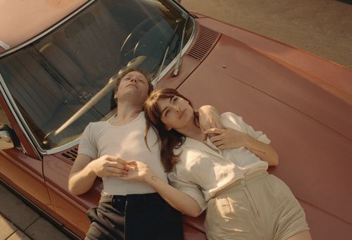 A woman and a man lie on the hood of a salmon-colored vintage car in the sunshine, with the woman resting her head on the man's arm.