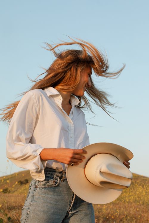 Blonde woman in a white shirt and jeans, holding a cowboy hat in a field with a small hill under a clear blue sky.