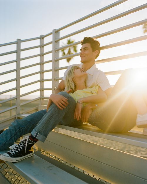 Young couple relaxes on bleachers in golden light. The woman leans against the man as they smile and enjoy the sunny outdoor setting.