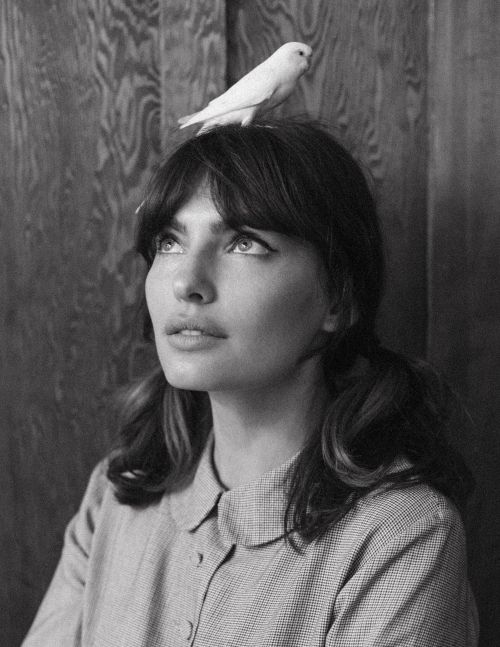 Monochrome portrait of a young woman with pigtails looking up with a white bird perched on her head in an indoor setting.