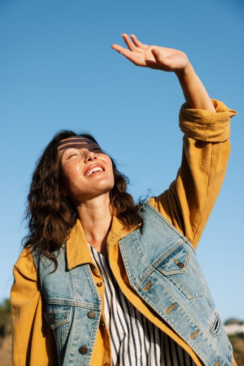 A woman in a denim and yellow jacket smiles, shielding her face from sunlight, set against a bright blue sky in an outdoor scene.