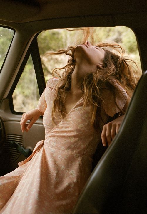 Young woman with long hair sits inside a vintage truck wearing a floral dress, head tilted back, and eyes closed. Trees are visible outside.