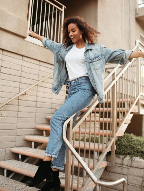 A smiling woman in a denim jacket and jeans walks down outdoor steps, balancing on the railing in sunshine. Casual fashion concept.