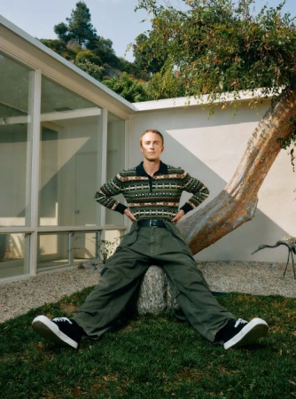Young man in patterned sweater and baggy green pants poses outside modern glass house, relaxed pose, natural light.