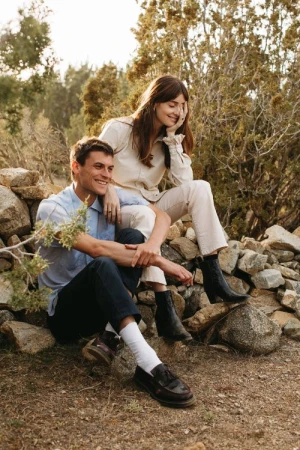 A smiling young man and woman sitting outdoors together amidst natural rock formations and dry brush vegetation.
