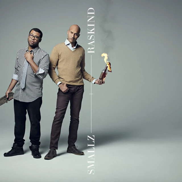 Two men stand in a studio, one holding a Molotov cocktail, the other a piece of wood. Vertical text divides the image.