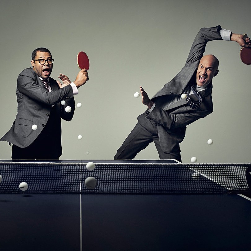 Two businessmen in suits playing table tennis, one with mouth open mid-swing, the other looks shocked, indoors on a dark table.