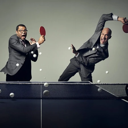 Two businessmen in suits playing table tennis, one with mouth open mid-swing, the other looks shocked, indoors on a dark table.