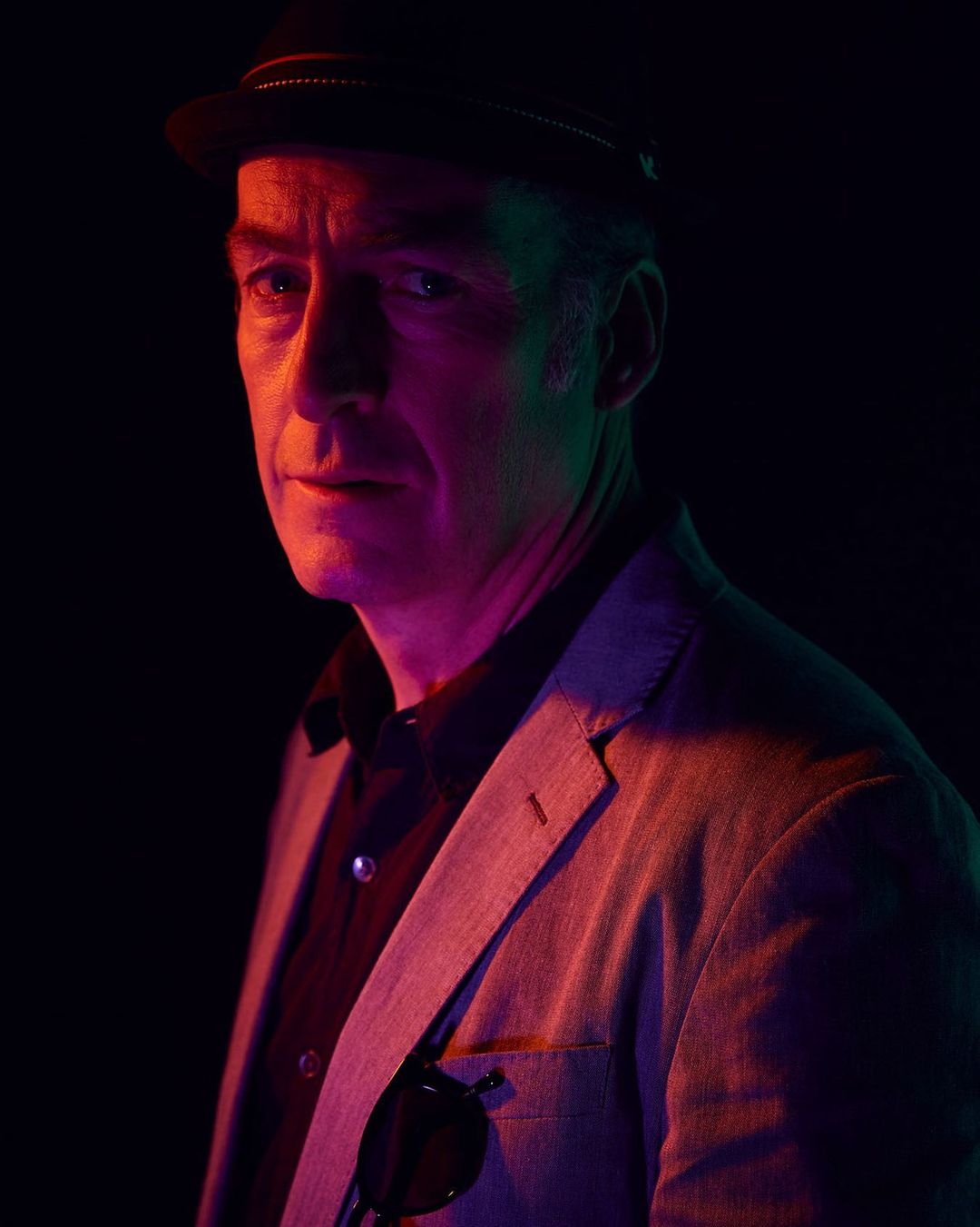 Portrait of a mature adult male wearing a hat and blazer in a dimly lit studio setting with colored lighting, striking a confident pose.