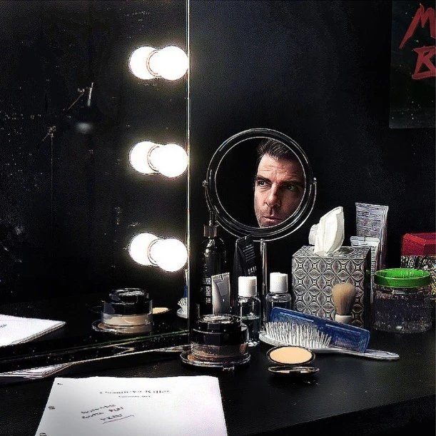 Reflection of a man's face in a round mirror surrounded by vanity lights, accompanied by an array of cosmetic products and papers.