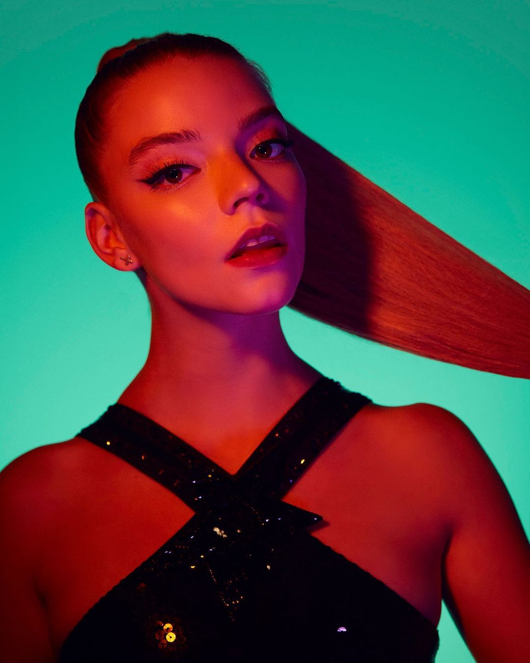 Portrait of a young adult female with makeup and a black halter top, posing against a teal backdrop with red lighting and her hair up.