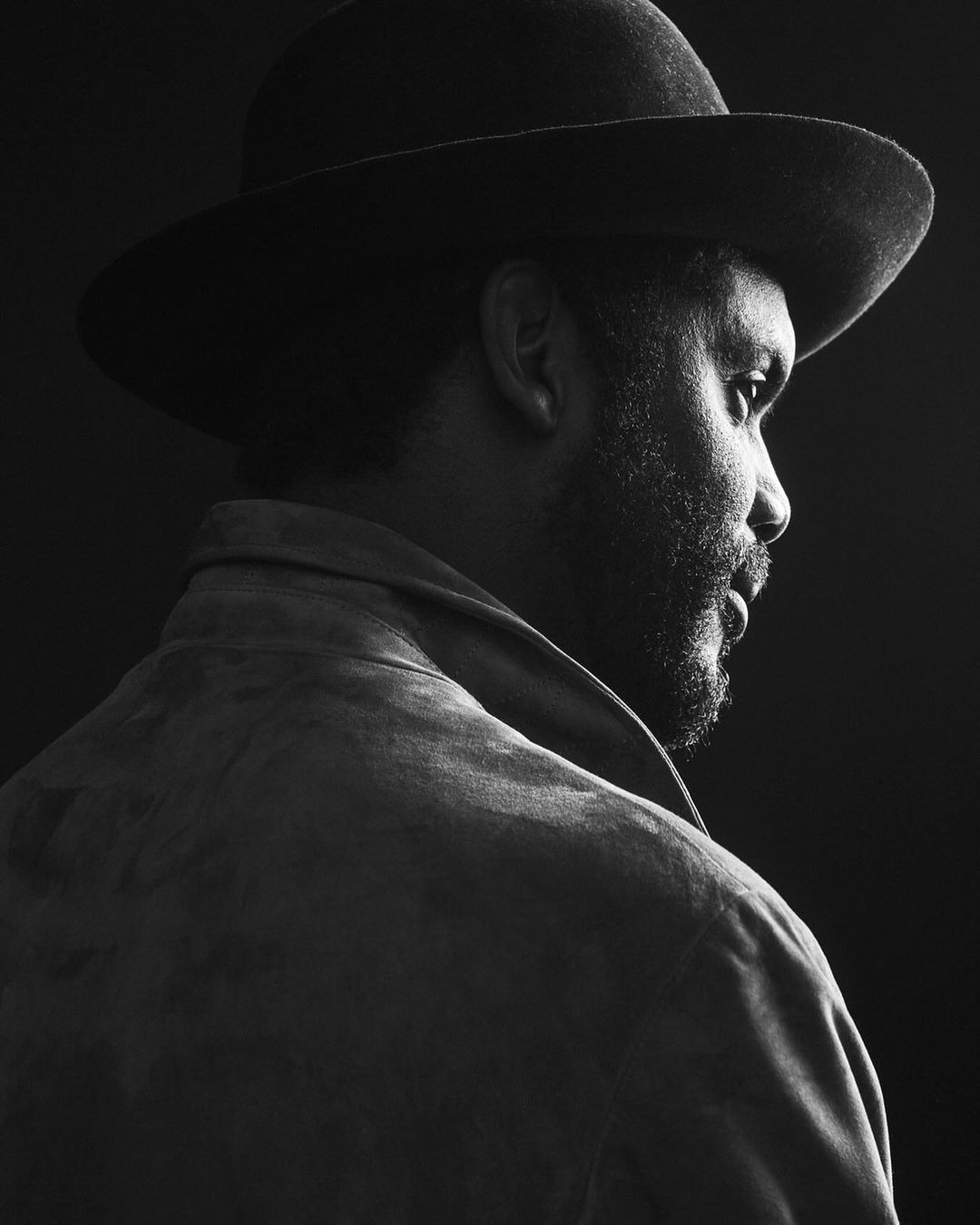 Black and white profile portrait of a man wearing a wide-brimmed hat and jacket against a dark background.