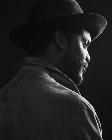 Black and white profile portrait of a man wearing a wide-brimmed hat and jacket against a dark background.