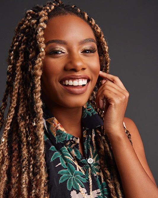 Close-up portrait of a smiling African-American woman with blonde braided hair wearing a black tropical print shirt against a grey backdrop.