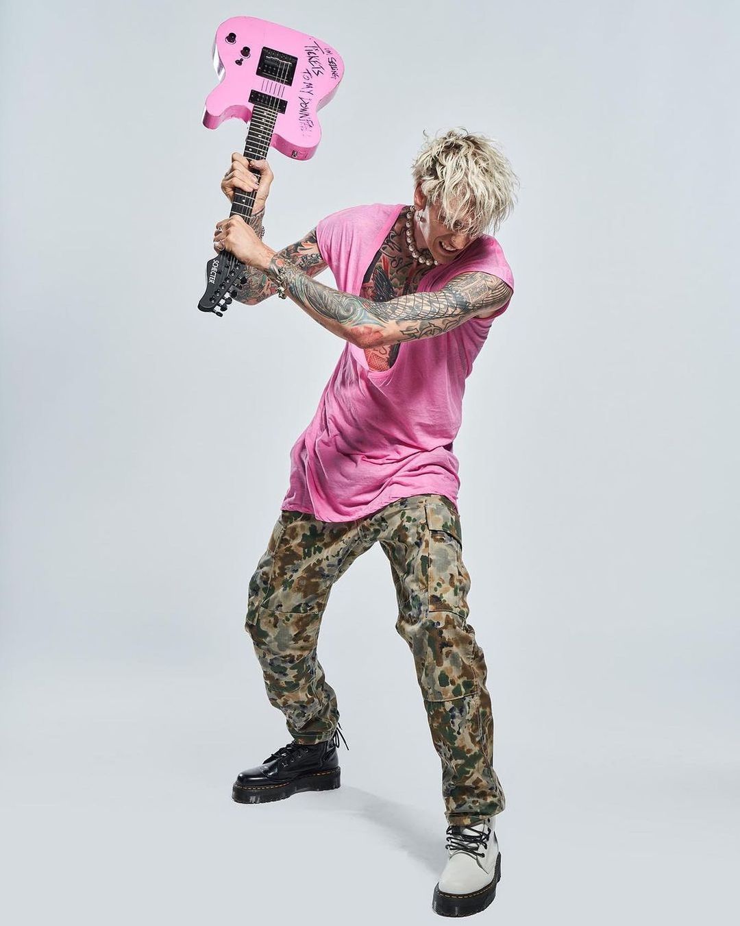 Tattooed man in pink shirt and camouflage pants poses with a pink electric guitar in a full shot against a white backdrop.