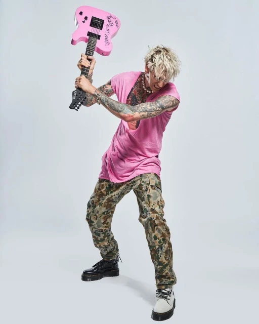 Tattooed man in pink shirt and camouflage pants poses with a pink electric guitar in a full shot against a white backdrop.