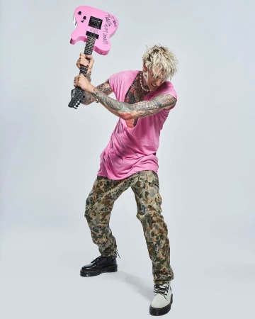 Tattooed man in pink shirt and camouflage pants poses with a pink electric guitar in a full shot against a white backdrop.