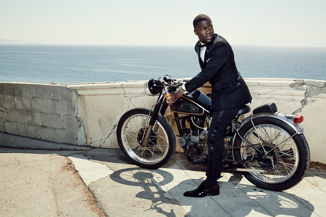 Adult male in a tuxedo poses with a motorcycle on a sunny day. Ocean and land are visible in the distance.