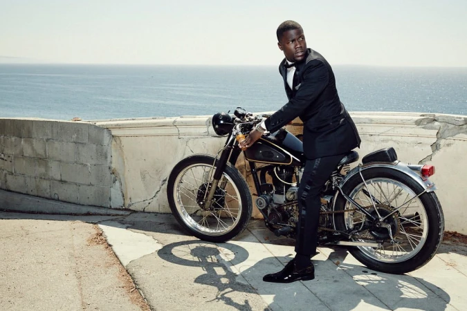 Adult male in a tuxedo poses with a motorcycle on a sunny day. Ocean and land are visible in the distance.