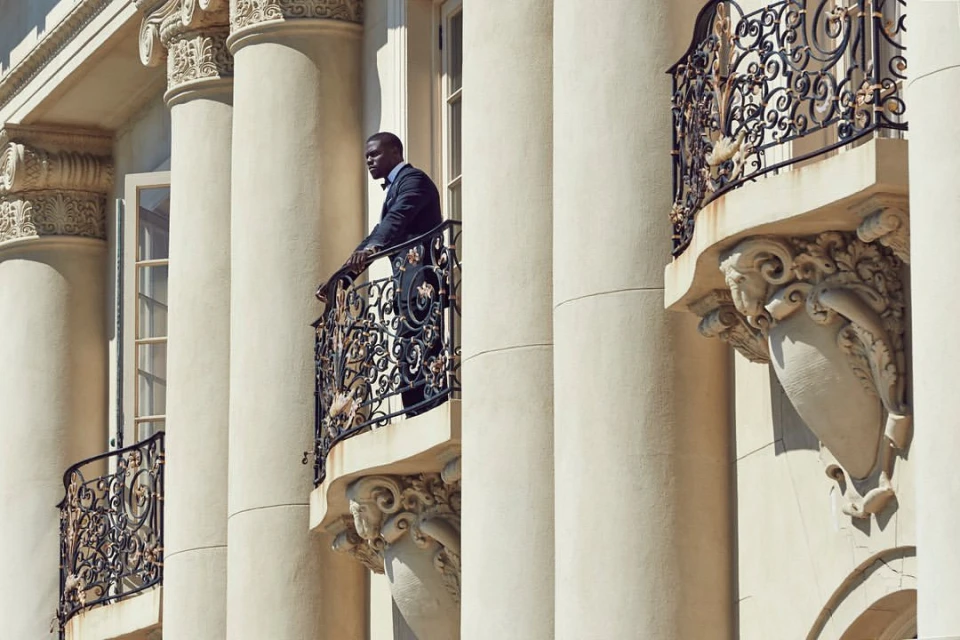 Elegant African American man in a suit standing on a balcony with ornate iron railings, surrounded by classical architectural columns.