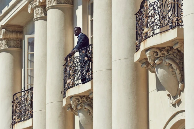 Elegant African American man in a suit standing on a balcony with ornate iron railings, surrounded by classical architectural columns.